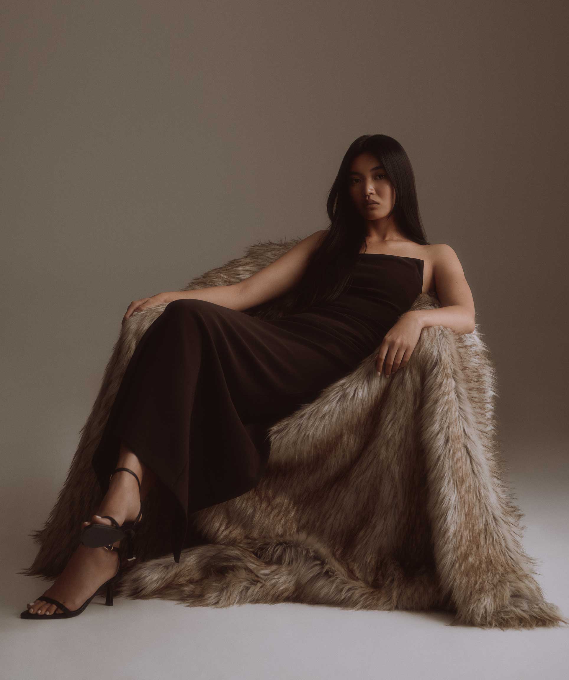 A woman with long dark hair sits draped in a shaggy taupe faux fur coat, wearing a dark brown strapless maxi dress and black strappy high-heeled sandals against a warm beige studio background.