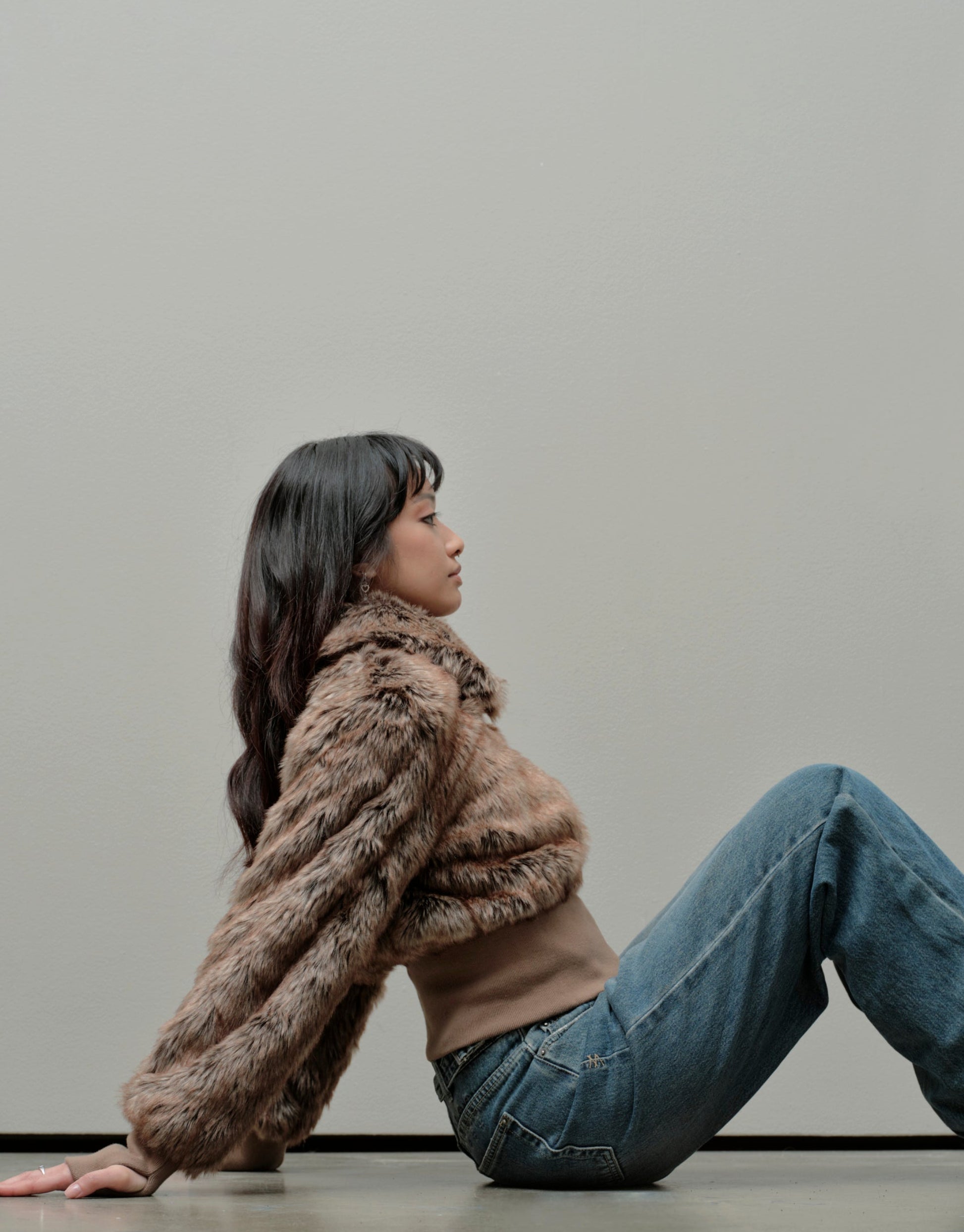 Person sitting on the floor wearing a brown faux fur coat and blue jeans against a plain background