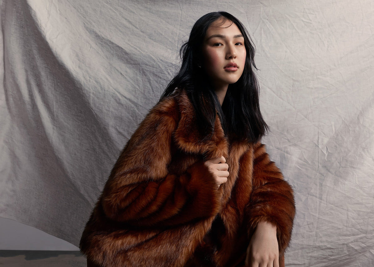 Woman wearing a brown faux fur coat and pants kneeling on a textured floor with a white draped fabric background.