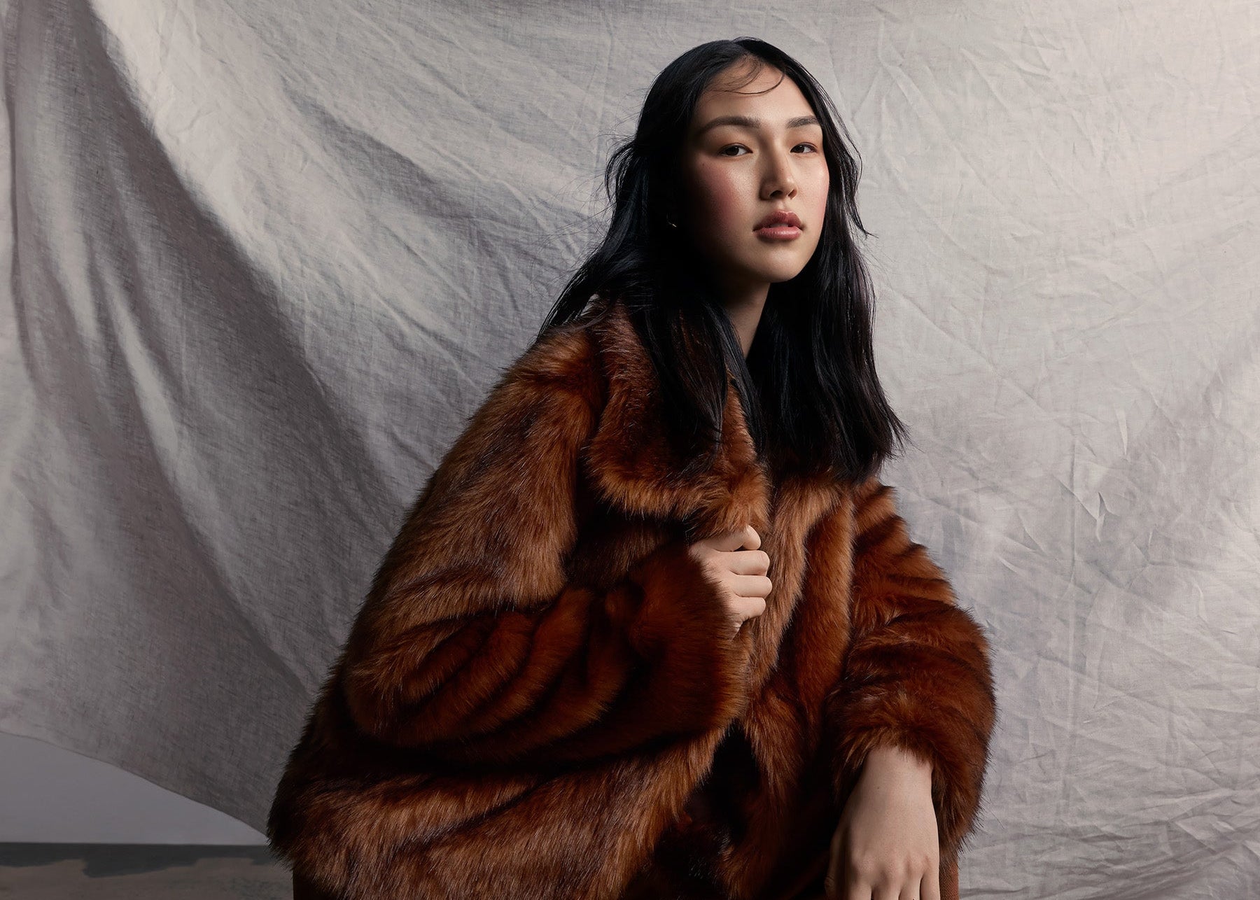 Woman wearing a brown faux fur coat and pants kneeling on a textured floor with a white draped fabric background.