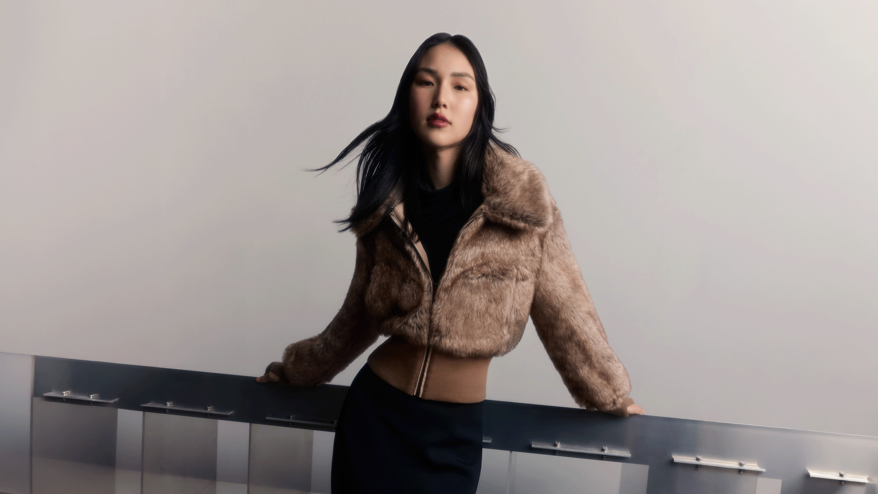A woman posing in a cropped, brown faux-fur jacket over a black top and black skirt, leaning on a glass railing against a neutral light gray background.