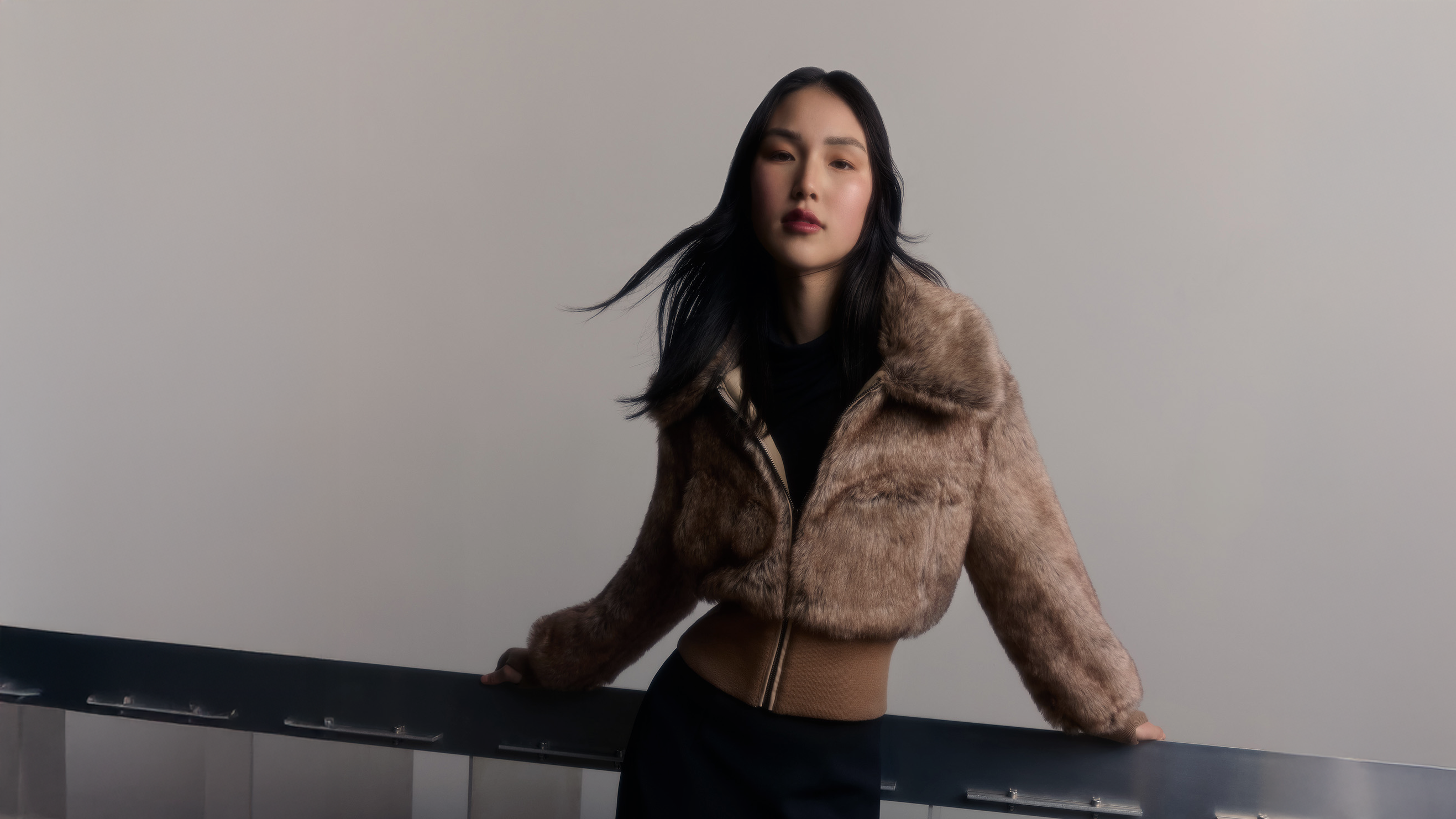A woman wearing a cropped, textured brown faux-fur jacket over a black top and black skirt, leaning on a dark railing against a plain light gray wall.