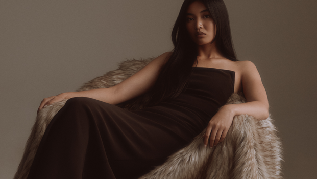 Woman in a dark dress sitting on a faux fur chair against a plain background